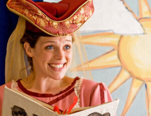 A woman is smiling and holding an open book in front of a painted backdrop featuring a sunshine