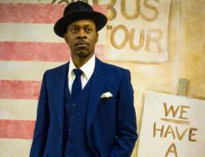 An actor portraying Dr Martin Luther King, Jr. stands in front of a painted backdrop depicting the American flag next to protest signs. He is wearing a navy blue suit, a tie, and a dark hat. 